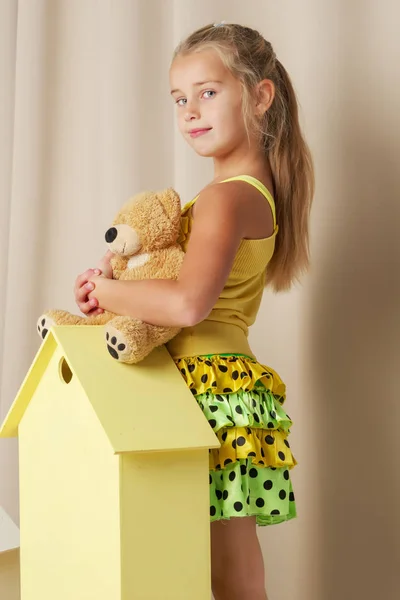 Menina brincando com um urso de pelúcia em uma casa de madeira de brinquedo . — Fotografia de Stock