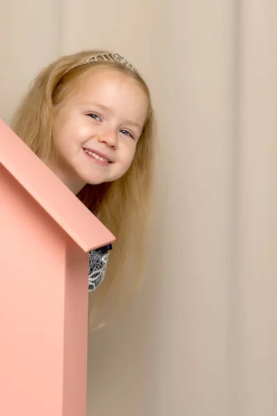 Een meisje kijkt uit achter een houten huis van speelgoed. — Stockfoto