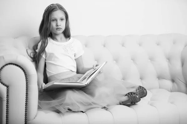 Ένα κοριτσάκι κάθεται στον καναπέ και διαβάζει ένα βιβλίο.. — Φωτογραφία Αρχείου