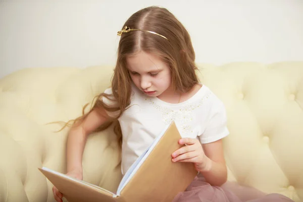 En liten flicka sitter på soffan och läser en bok.. — Stockfoto