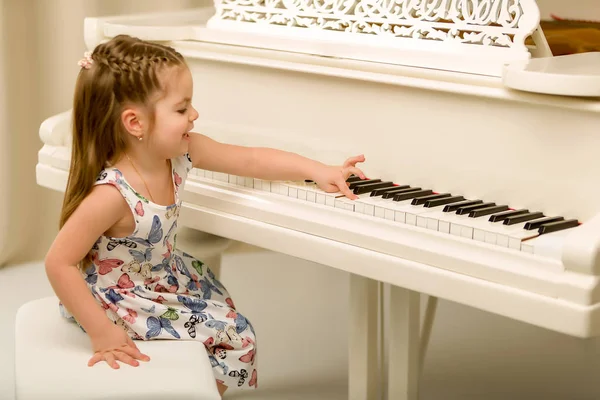 En vacker liten flicka spelar på en vit flygel. — Stockfoto