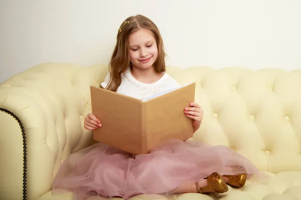 Uma menina está sentada no sofá e lendo um livro. — Fotografia de Stock