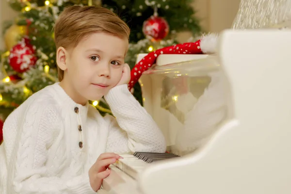 Um menino está sentado ao piano perto da árvore de Natal. — Fotografia de Stock