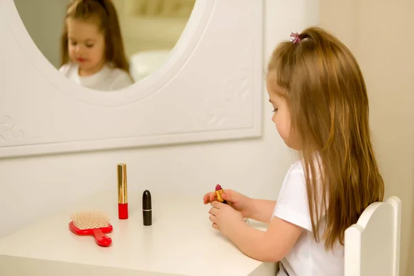 A little girl near the mirror paints lipstick lips — Stock Photo, Image
