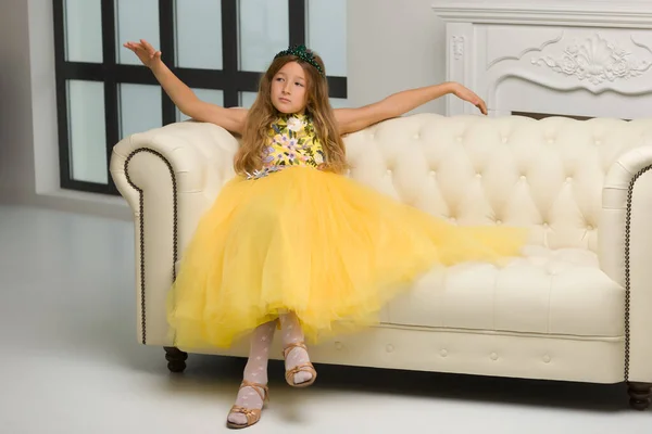 Uma menina pequena e elegante senta-se em um sofá branco perto de um grande vento — Fotografia de Stock