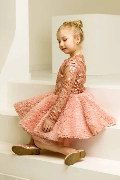 Jolie petite fille est assise dans le studio sur les escaliers blancs. style et concept de mode, enfance heureuse. — Photo