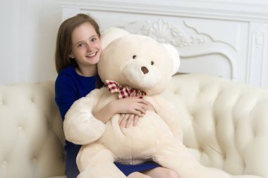 Okul çağındaki güzel genç kız elinde oyuncak ayıyla kanepede oturuyor..