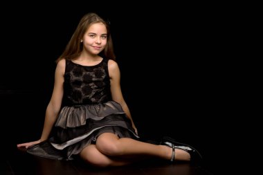 Küçük bir kız stüdyoda yerde, siyah bir arka planda oturuyor..