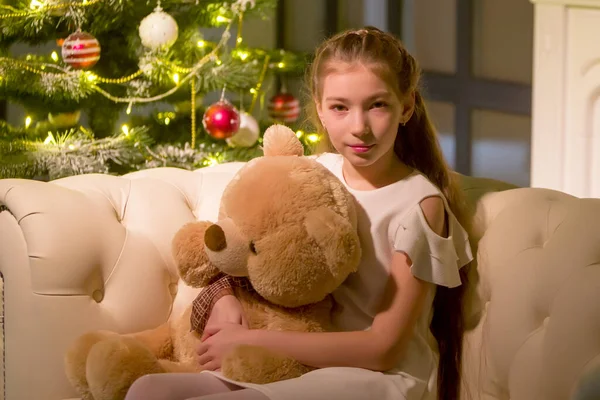 Oyuncak ayılı küçük bir kız Noel ağacının yanındaki kanepede oturuyor.. — Stok fotoğraf