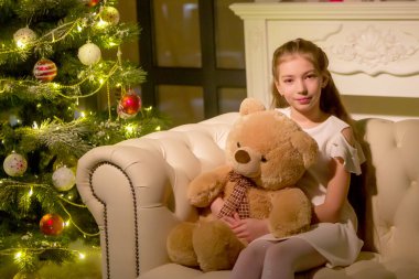 Oyuncak ayılı küçük bir kız Noel ağacının yanındaki kanepede oturuyor..