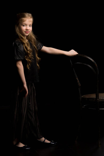 Portrait of a girl standing on a black background near the old Viennese chair.