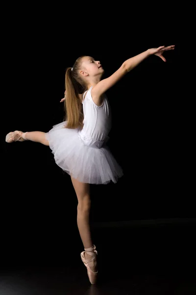 Schattig klein meisje in een tutu en pointe schoenen is dansen in de studio op een zwarte achtergrond. — Stockfoto