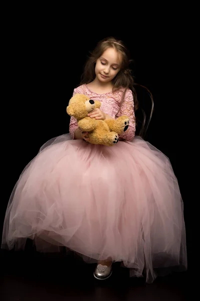 Krásná dívka v pěkné růžové šaty objímající medvídek s něžností. — Stock fotografie