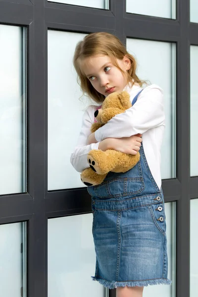 A little girl stands by the window and hugs a teddy bear.