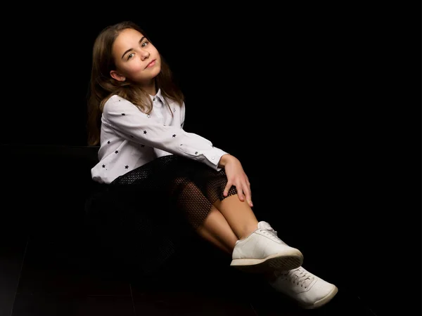 Een klein meisje zit in de studio op de vloer op een zwarte achtergrond. — Stockfoto