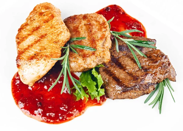 Kızarmış ızgara sığır eti, kümes hayvanları ve kırmızı yaban mersini sosuyla süslenmiş domuz bifteği ve beyaz arka planda izole edilmiş otlar.