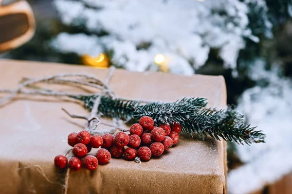 Dekoratif el yapımı kış bayramı hediye paketi ladin dalı, kırmızı böğürtlen ve arka planda yapay karla kaplı Noel ağacı