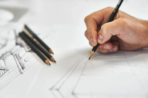 Graphic designer and artist drawing architectural blueprint with his right hand, several black pencils in background on white white sheet of paper