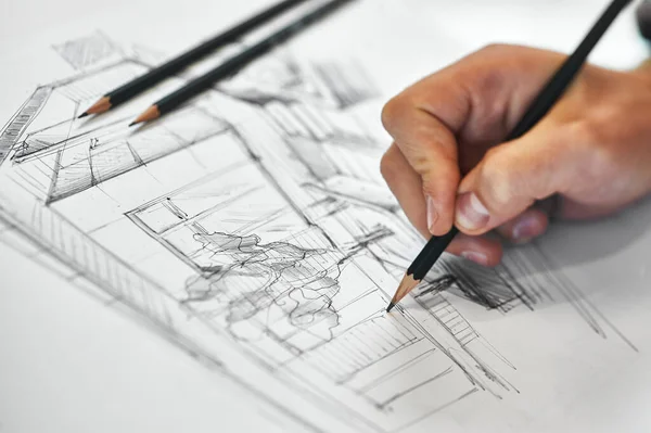 Graphic designer and artist drawing architectural blueprint with his right hand, several black pencils in background on white white sheet of paper