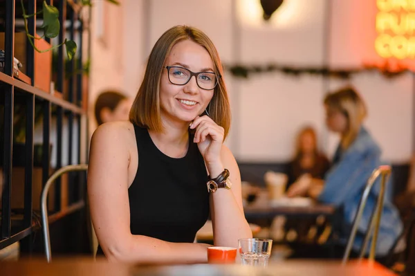 Linda chica con una hermosa sonrisa en gafas sentadas en un café. Tomando café, sosteniendo una tapa de café —  Fotos de Stock