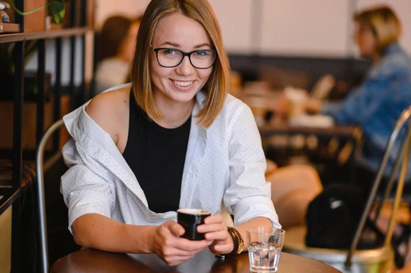 Linda chica con una hermosa sonrisa en gafas sentadas en un café. Tomando café, sosteniendo una tapa de café —  Fotos de Stock