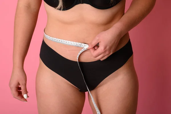 young woman size plus measuring her waist with a tape measure on pink background