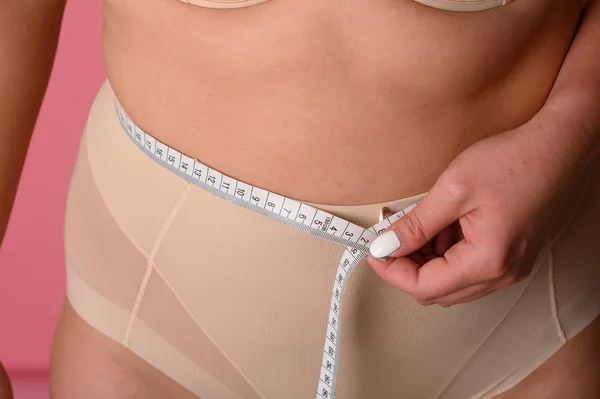 Young woman size plus measuring her waist with a tape measure on pink background — Stock Photo, Image