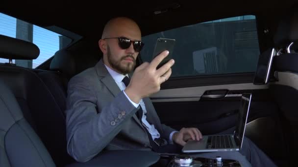 Jóképű üzletember használja a mobilját egy modern autóban, egy sofőrrel a város közepén. Fogalom az üzleti, siker, utazás, luxus — Stock videók