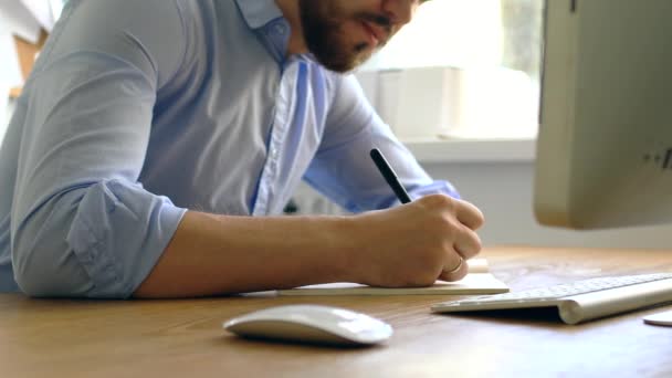 En man i blå skjorta antecknar i en anteckningsbok. Närbild: mannen böjer sig över skrivbordet och gör anteckningar med en penna, lutande på bordet. — Stockvideo