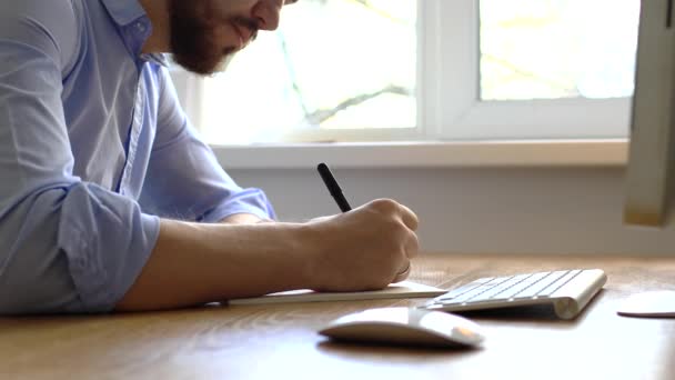 Un hombre con una camisa azul hace una nota en un cuaderno. Primer plano: el hombre se inclina sobre el escritorio y toma notas con un bolígrafo, apoyado sobre la mesa . — Vídeos de Stock