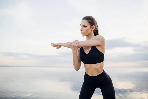 Mujer joven en ropa deportiva negra estirando las manos junto al río. Chica de fitness con las manos levantadas por encima de su cabeza estirándose al aire libre en el soleado día de verano — Foto de Stock