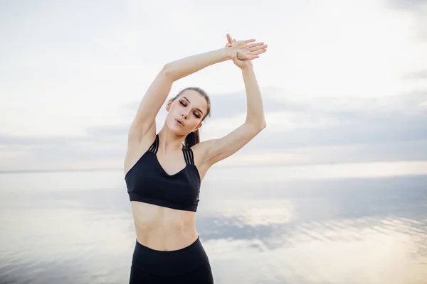 Siyah spor kıyafetli genç bir kadın nehrin kenarında ellerini uzatıyor. Güneşli bir yaz gününde ellerini başının üstüne kaldırmış spor yapan bir kız. — Stok fotoğraf