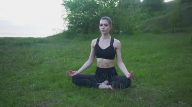 Spor elbiseli genç bir kız parkta yoga yapıyor. Kız Lotus pozisyonunda oturuyor.