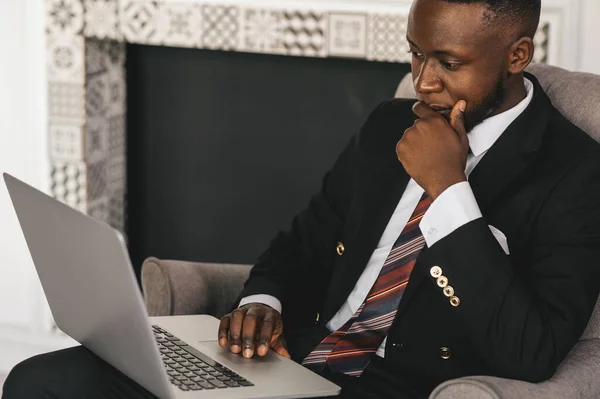 Bonito empresário afro-americano em terno clássico está usando um laptop e sorrindo enquanto trabalha no escritório moderno — Fotografia de Stock