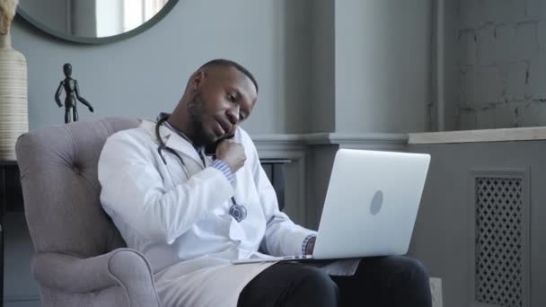 Un jeune médecin afro-américain travaille sur un ordinateur portable et parle au téléphone. Le docteur a beaucoup de travail à faire. — Video