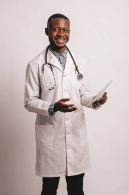 Siyah tenli genç doktor elinde kağıtlarla beyaz bir arkaplanda gülümsüyor.