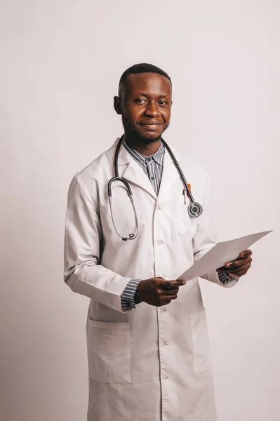 Czarny młody uśmiechnięty lekarz stoi na białym tle z papierami w rękach — Zdjęcie stockowe