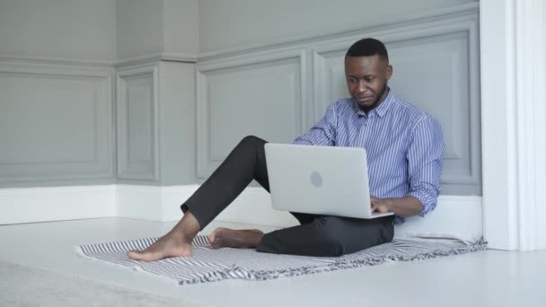 Молодий чорний чоловік працює на ноутбуці вдома, сидячи на підлозі. робота або навчання вдома — стокове відео