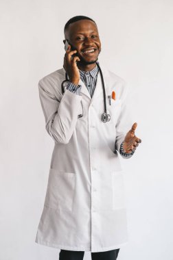  Siyah tenli genç doktor gülümsüyor. Beyaz bir arkaplanda telefonda konuşuyor.