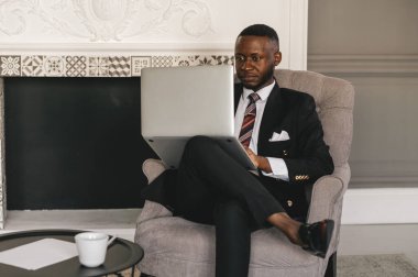 Siyah takım elbiseli siyah bir adam elinde dizüstü bilgisayarla sandalyede oturuyor.