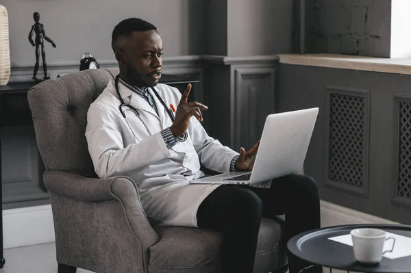 dark-skinned doctor talking on skype zoom with laptop waving