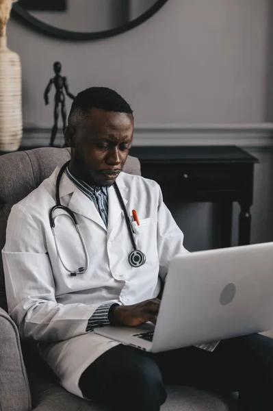 Czarny lekarz na szarym krześle z laptopem zdalnie konsultuje się z pacjentem — Zdjęcie stockowe