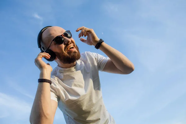Portrét pohledného mladého muže s vousy poslech hudby se sluchátky proti zakalené obloze pozadí — Stock fotografie