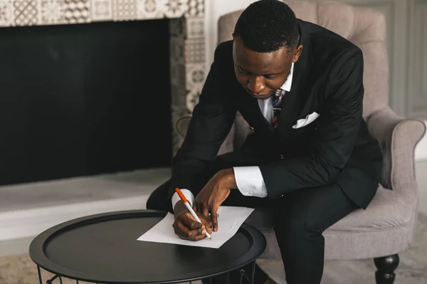 Empresário americano africano em um terno faz anotações em uma folha de papel enquanto sentado em seu escritório perto da lareira — Fotografia de Stock