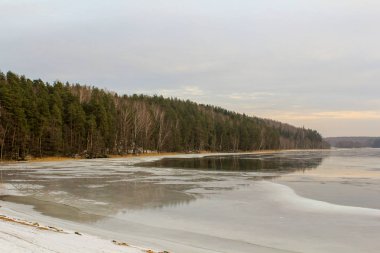 Tepelere kıyı ve buz kenar boyunca kapsayan orman ve göl günbatımı gri gökyüzü ile yüzeyi yansıyan