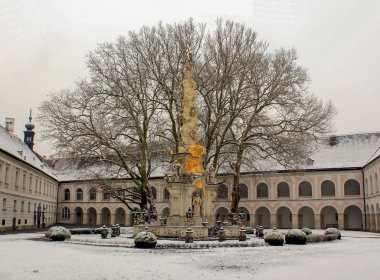 Sütunlar ile bir iki katlı pembe bina ve kemerler ile eski bir çıplak ağaca ve altındaki beyaz ve altın anıt avlusu