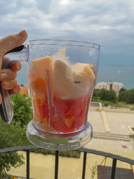 Büyük kesim meyve parçaları ve mavi deniz ve gri gökyüzüne karşı bir kadının elinde dondurma birkaç kaşık ile blender kase