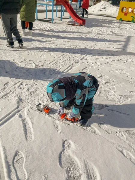冬のオーバーオールの小さな男の子は 雪の中でおもちゃのオレンジ色のトラクターとダンプトラックで遊びます — ストック写真