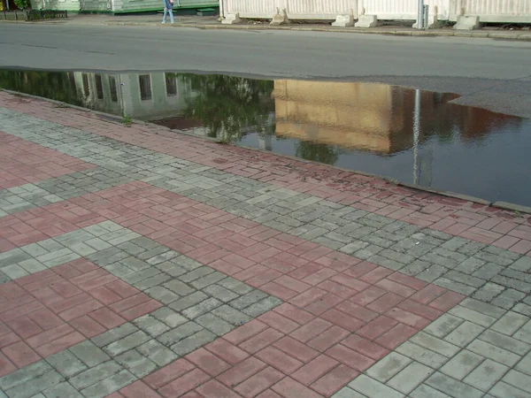 A puddle on the road next to a sidewalk paved with tiles and reflecting multicolored houses in it