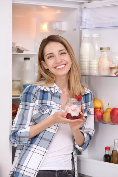 Young attractive woman eating tasty yogurt near fridge